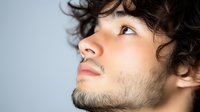 Young Caucasian male model with curly dark hair and facial stubble looking up thoughtfully against light background in profile view portrait showing contemplative expression.