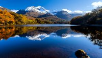 Snow-capped mountain peaks reflect in calm lake waters during autumn, with golden foliage framing dramatic landscape and pristine wilderness under blue sky.