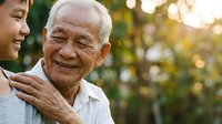 Senior Asian man with white hair and glasses smiling warmly while embraced by younger woman outdoors in garden setting during golden hour, representing family bonding and intergenerational love.
