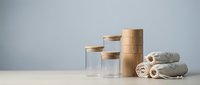 Minimalist bathroom accessories with glass jars, bamboo lids and rolled towels on white surface against gray wall for spa-inspired home decor.