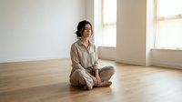 Young Asian woman in beige linen pajamas sitting cross-legged on wooden floor in bright minimalist room practicing morning meditation and mindfulness routine.
