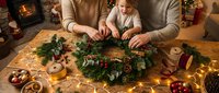 Mother and toddler daughter making Christmas wreath together at wooden table with festive decorations, fireplace glowing in cozy living room background.