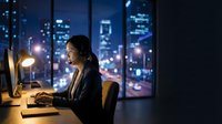 Professional businesswoman working late at computer in modern office with city skyline view through windows at night for corporate productivity concepts.