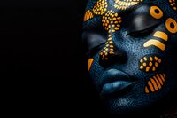 African woman with creative blue and gold face paint featuring tribal patterns and dots against dark background, artistic beauty portrait.
