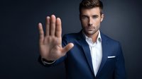 Confident businessman in navy blue suit making stop gesture with palm forward against dark background, conveying authority and boundaries.