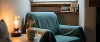 Cozy reading nook with teal armchair, warm lamp lighting, wooden shelves with books, and steaming mug on side table. Perfect for relaxation and home interior design projects.