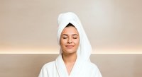 Woman with wrapped white towel on head wearing bathrobe after shower or spa treatment, smiling at camera against neutral beige background for wellness and beauty content.