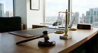 Professional law office interior with wooden gavel and brass scales of justice on desk overlooking modern city skyline through large windows.