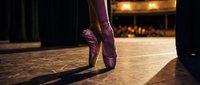 Ballerina standing on pointe in pink satin ballet shoes on a dramatic stage with warm spotlight and blurred audience in background.