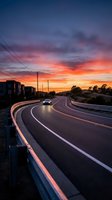 Curved highway with vehicle traveling during vibrant sunset, dramatic orange and purple sky, long exposure light trails on road markings, ideal for transportation and travel concepts.
