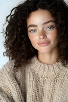 Young Mediterranean woman with natural curly dark hair and blue eyes wearing cream knit sweater smiling at camera against light background. Soft beauty portrait.