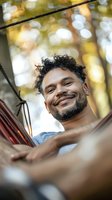Mixed race man with curly hair relaxing in hammock outdoors, genuine smile and casual attitude against blurred natural bokeh background. Wellness and lifestyle concept.