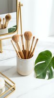 Wooden makeup brushes in ceramic holder on white marble vanity with gold mirror and tropical plant for beauty routine organization.