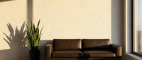 Brown leather sofa positioned against beige wall with potted plant and natural sunlight casting shadows, suitable for interior design and home decor projects.