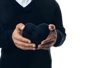 Male hands holding black heart shape against white background, wearing dark sweater. Minimalist composition expressing love, care and emotional connection.