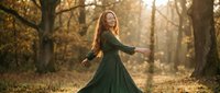 Young Caucasian woman with long red hair spinning in flowing green dress during golden hour in autumn forest with warm sunlight filtering through trees.