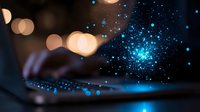 Glowing blue particles float above laptop keyboard with blurred warm bokeh lights in dark background creating magical digital atmosphere.