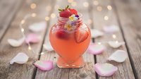 Refreshing strawberry cocktail in glass jar with rose petals on rustic wooden table with warm bokeh lights creating romantic atmosphere for celebrations.