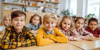 Diverse group of elementary school children sitting at wooden desk in classroom, smiling at camera with bookshelf in background.