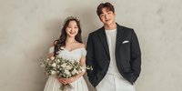 Young Asian bride in white wedding dress with floral crown and bouquet standing beside groom in black suit jacket against neutral background.