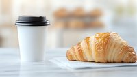 Fresh baked croissant served on white napkin with takeaway coffee cup on marble table, morning breakfast setting with soft natural lighting.
