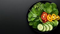 Fresh vegetable bowl with sliced cucumber, bell peppers, spinach leaves on black plate against dark background, top view. Food photography for healthy eating.