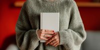 Person in oversized gray knit sweater holding blank white book against red background, showcasing cozy autumn or winter reading atmosphere.