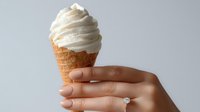 Creamy soft serve vanilla ice cream in waffle cone held by female hand with engagement ring against neutral gray background.