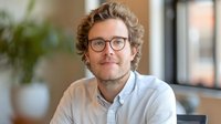 Young Caucasian male professional with curly hair and glasses smiling at camera in modern office setting. Natural lighting creates warm atmosphere for business portrait.