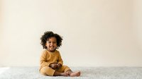 Young child with curly dark hair and medium skin tone wearing mustard yellow clothing, sitting on gray carpet against neutral beige wall background for lifestyle and family content.