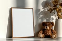 Brown teddy bear sitting next to wooden frame mockup and white ceramic vase with dried flowers on table, natural shadow play on wall, minimalist home decor.