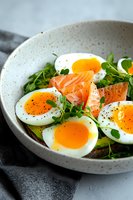Soft boiled eggs with smoked salmon, fresh microgreens and avocado served on dark bread in ceramic bowl. Black pepper seasoning adds finishing touch.