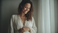 Young woman with wavy brown hair smiling by window in white cotton blouse, soft natural light creating intimate morning atmosphere.