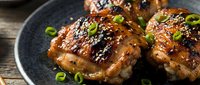Grilled chicken thighs with teriyaki glaze and green onions on dark plate. Asian cuisine dish with caramelized sauce for restaurant menu and cooking content.