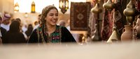 Young Middle Eastern woman in traditional black embroidered dress smiling warmly at bustling marketplace with ornate brass items and cultural artifacts.