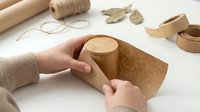 Hands crafting wooden cylindrical container with natural cork lid on white workspace surrounded by eco-friendly packaging materials and twine.