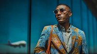 Young African American businessman in stylish patterned suit at airport terminal with airplane view through window, professional travel concept.