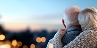 Senior couple embracing while watching sunset over city lights, rear view showing white hair and warm sweaters, creating intimate romantic moment at dusk.