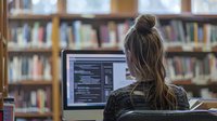 Young Caucasian woman programmer with messy bun hairstyle working on computer code in library setting, view from behind at screen displaying programming syntax.