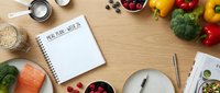 Overhead view of meal planning notebook surrounded by fresh vegetables, fruits, and cooking utensils on wooden surface for nutrition and diet planning projects.