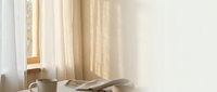 Cozy morning scene with soft sunlight streaming through sheer curtains onto unmade bed with white linens and coffee cup creating warm peaceful bedroom atmosphere.