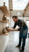 Male artisan potter in apron examining handmade ceramic vessels on wooden shelves in bright studio workshop with skylights, showcasing craftsmanship and pottery creation.
