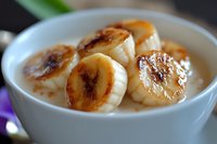 Caramelized banana slices floating in creamy yogurt or milk dessert, close-up view showing golden-brown surface and smooth texture in white ceramic bowl.