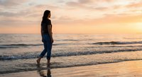 Young woman walking barefoot on beach at sunset with golden sky and ocean waves creating peaceful silhouette scene for wellness and travel concepts.