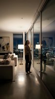 Woman in brown cardigan standing in modern high-rise apartment at night, gazing at city skyline through floor-to-ceiling windows. Contemporary interior design with minimalist furniture and ambient lighting for luxury lifestyle and real estate.