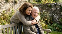 Middle-aged Caucasian woman embracing elderly Caucasian man on garden bench. Loving daughter hugging senior father outdoors. Family bonding moment in peaceful setting.