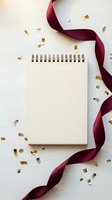 Blank spiral notepad with burgundy ribbon and gold confetti on white wooden background for celebration planning and party invitations.