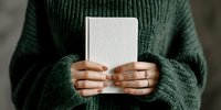 Person in green knitted sweater holding blank white notebook or journal with manicured nails, creating minimalist lifestyle concept.