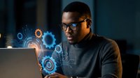 Young African American software engineer working with holographic interface technology displaying digital gears and circuits in modern office environment.