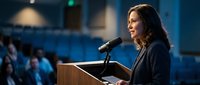 Professional businesswoman speaking at podium during corporate conference presentation with audience in modern venue.